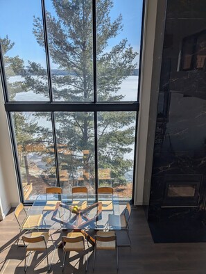 A dining area with a glass table and chairs, a fireplace, and a view of trees and water through large windows. - Beautiful Eco-Cottage near Parry Sound (The Archipelago)