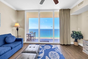 Ein Zimmer mit blauem Sofa, einem Tisch mit Lampe, einem gemusterten Teppich, einer Topfpflanze und Blick auf das Meer durch eine Schiebetür.