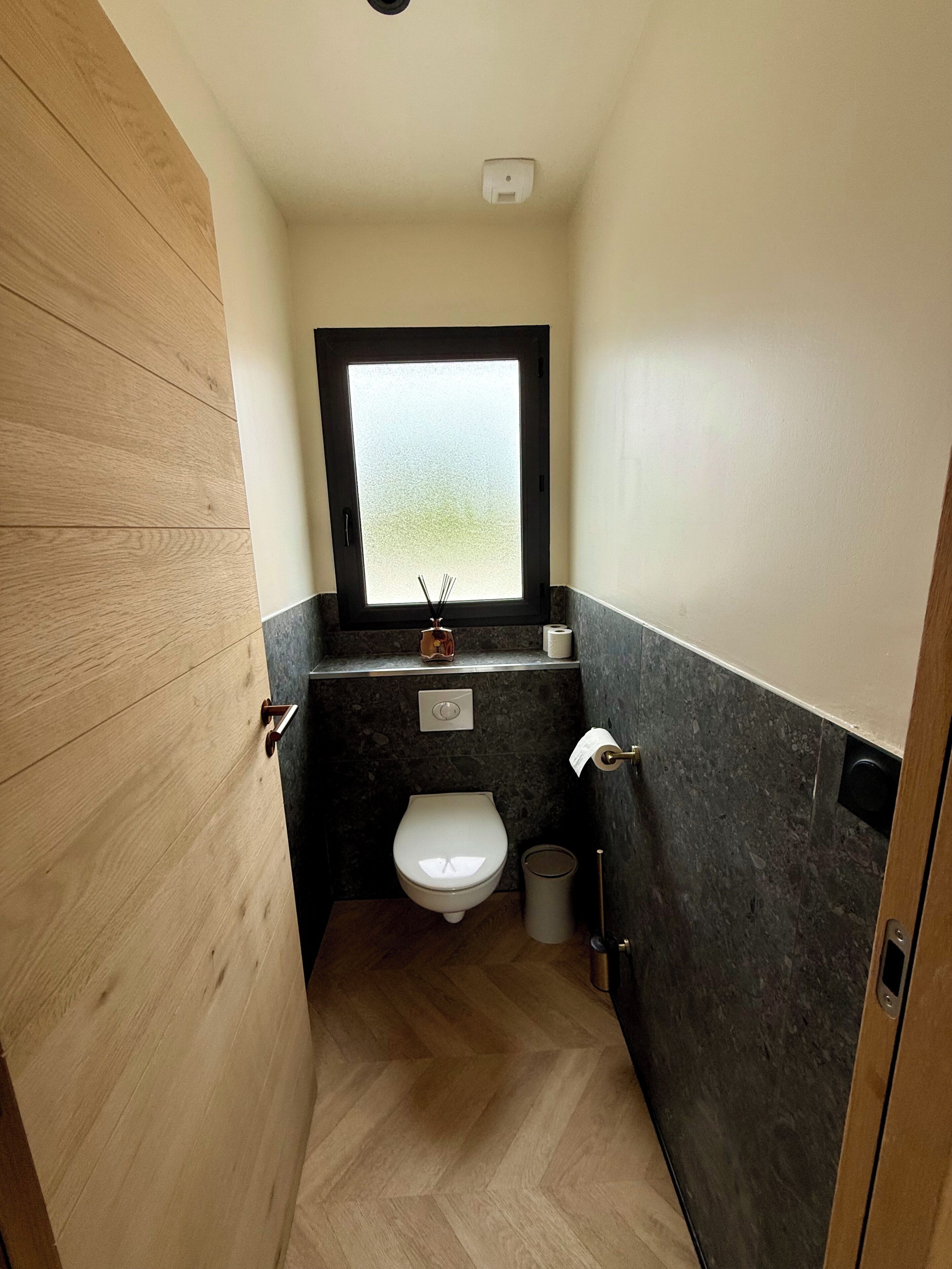A small bathroom with a toilet, a window, a sink, and a trash bin.