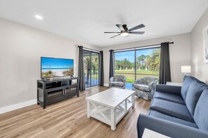 Ein modernes Wohnzimmer mit Flachbildfernseher, Deckenventilator, Sofa, Couchtisch und großen Fenstern mit Vorhängen.