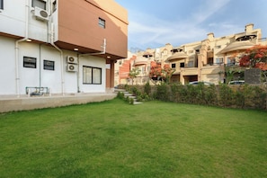 Um edifício residencial com telhado plano, um pequeno jardim frontal e céu claro.