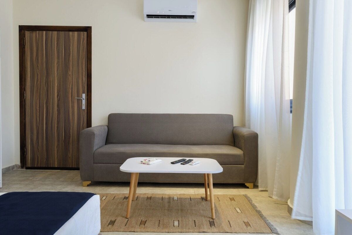 A room with a sofa, a small white table with remote controls, a wooden door, and an air conditioning unit on the wall.