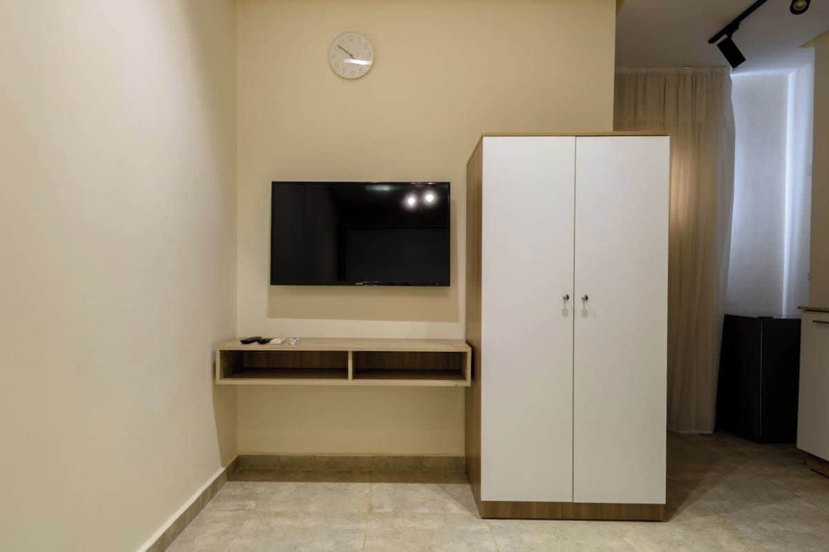 A room with a flat-screen TV mounted on the wall, a white wardrobe, and a small shelf below the TV.