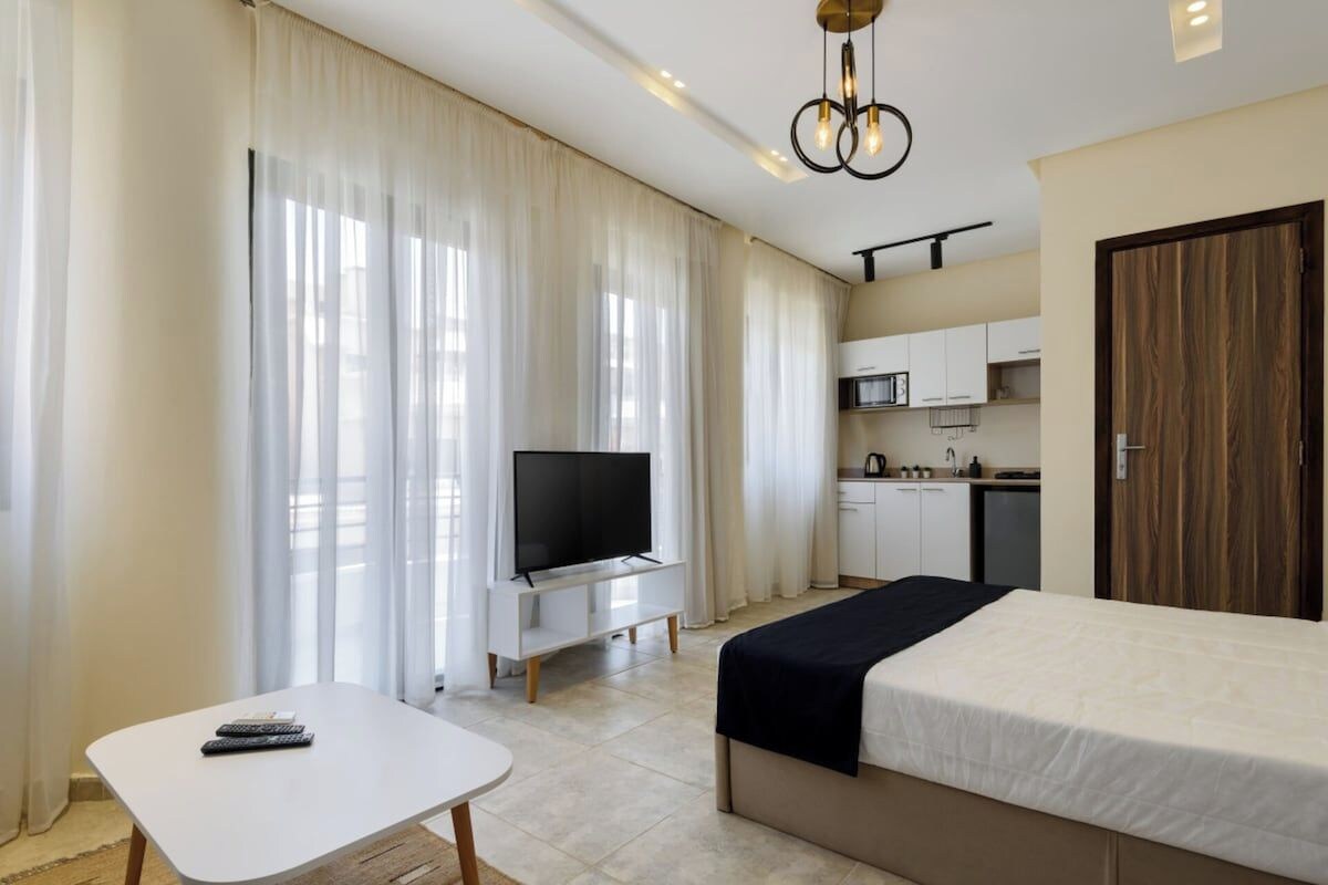 A hotel room with a bed, a TV, a small table, and a kitchenette in the background.