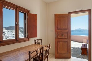 Une pièce avec une table et des chaises en bois, une grande fenêtre donnant sur une ville côtière, et une porte menant à un espace extérieur doté d’un sol en pierre.