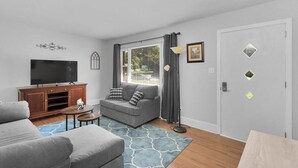 A living room with a grey sofa, a television on a wooden cabinet, a window with curtains, and a door with diamond-shaped stickers. - Cozy Pet Friendly Home with Private Backyard (Charlotte)