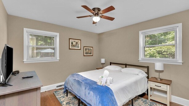 A bedroom with a bed, a desk with a television, a ceiling fan, and two windows with blinds. - Cozy Pet Friendly Home with Private Backyard (Charlotte)