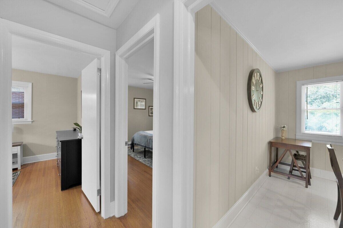 A hallway with a wooden floor, a desk with a chair, a clock on the wall, and a bed in the background.
