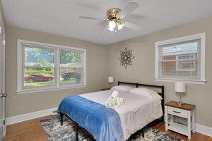 A bedroom with a bed, a ceiling fan, two windows, a nightstand, and a decorative wall piece. - Pet-Friendly 2BR | Fenced Yard & Near Plaza Midwood (Charlotte)