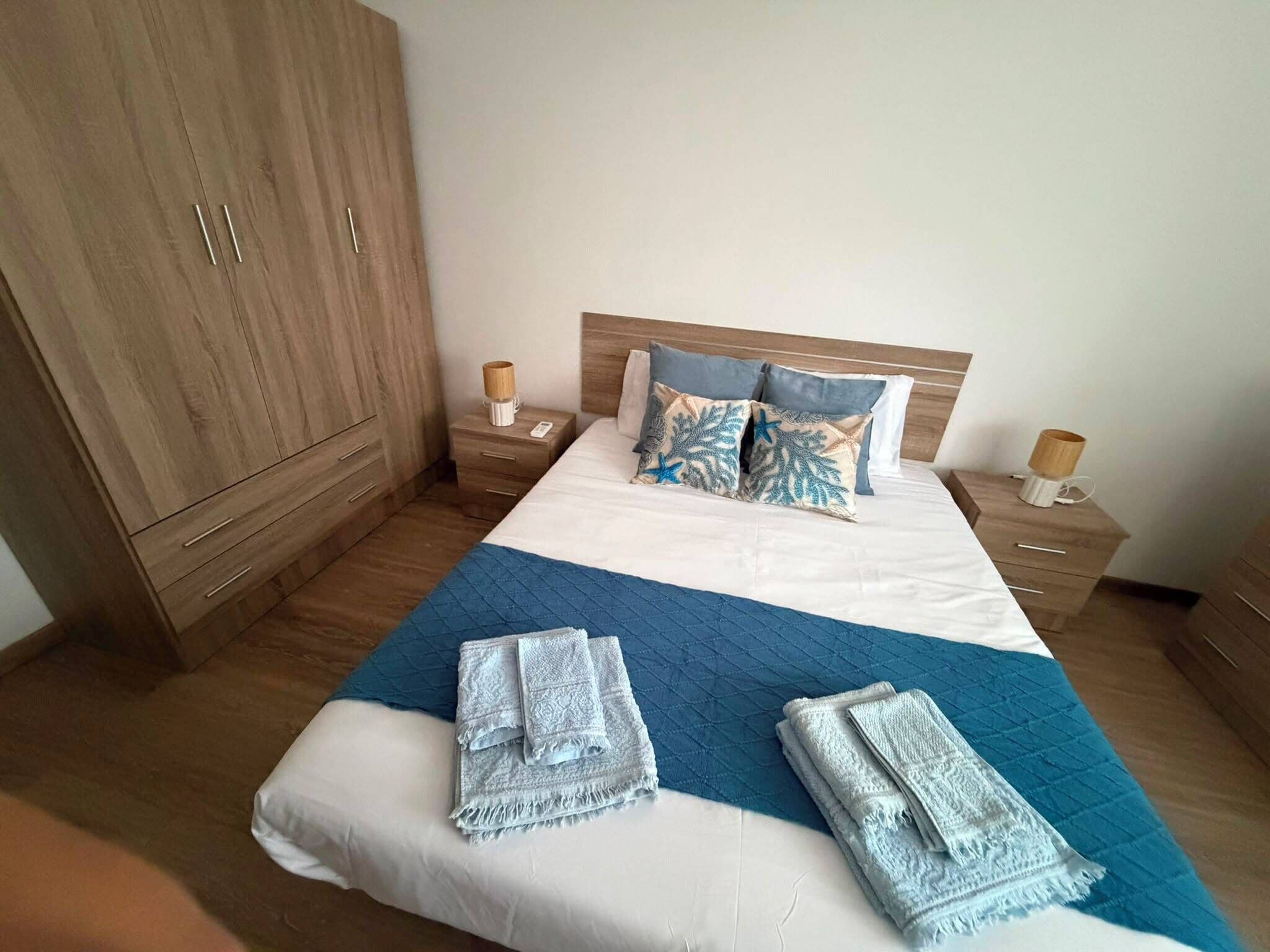 Habitación de hotel con una cama grande, armario de madera, mesitas de noche y colcha azul acolchada.
