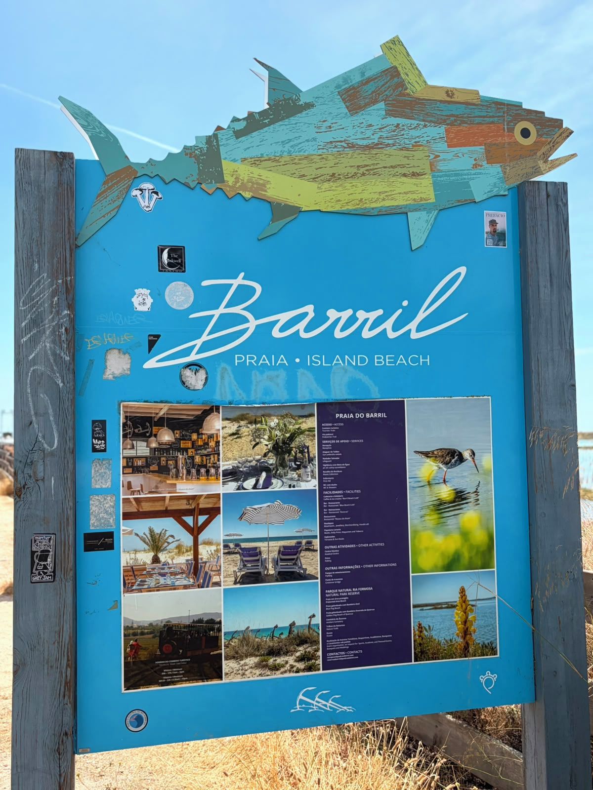 A beach sign with a fish design, the name 'Barril Praia - Island Beach', and various informational panels including photos of beach activities and amenities.