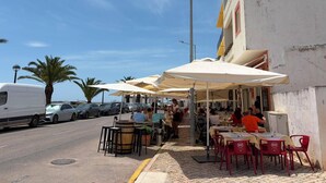 Un café con mesas al aire libre, una furgoneta blanca estacionada al lado y palmeras al fondo.