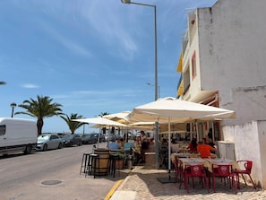 A street-side café with outdoor seating, a white van parked on the side, and palm trees in the background. - JR Santa Luzia (Tavira)