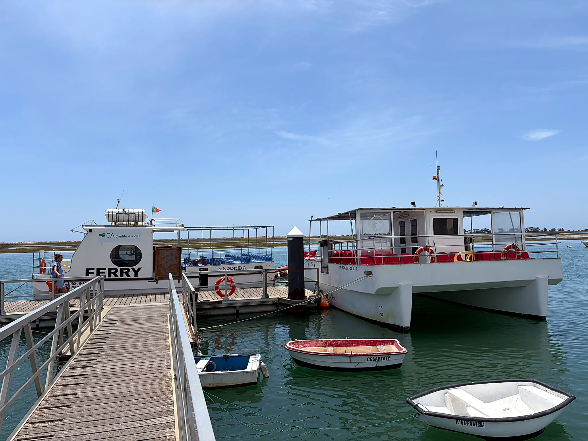 A ferry dock with a wooden pier, a white ferry with red seating, and two smaller boats.