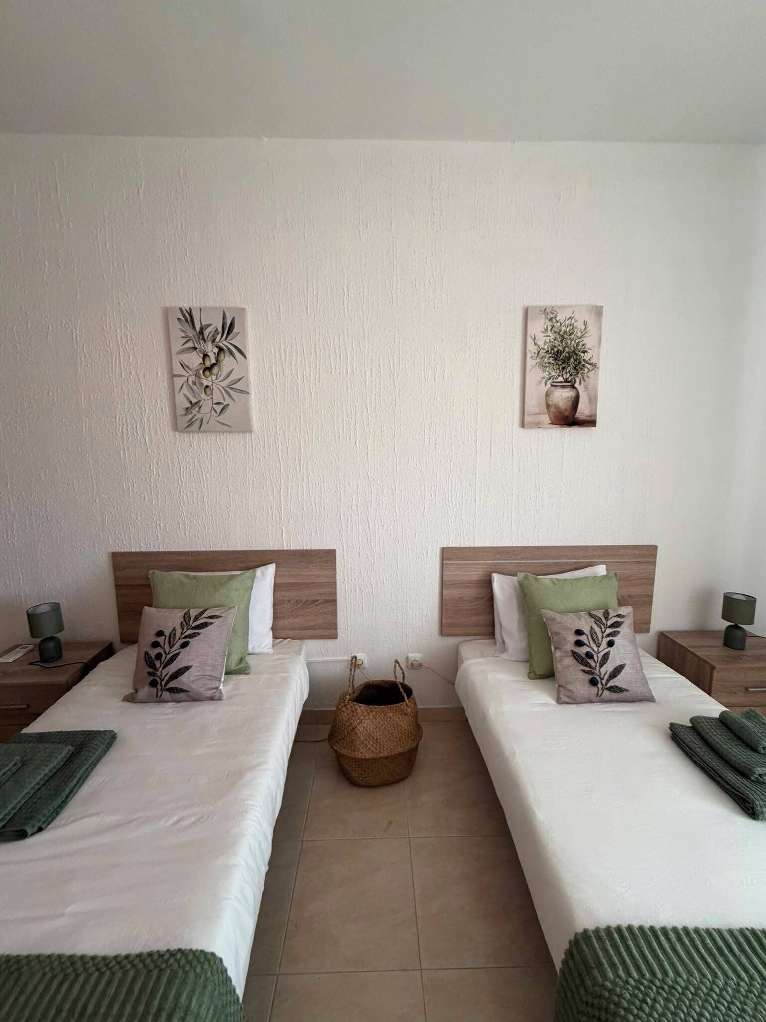 Habitación de hotel con dos camas, cada una con cabecero de madera, ropa de cama blanca y almohadones verdes. Dos cuadros en la pared, uno con una planta y otro con un jarrón de flores. Una cesta tejida está colocada entre las camas.