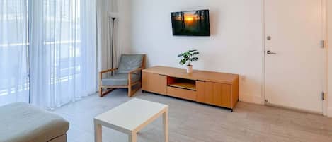Un salon moderne comprenant un canapé, une table basse, un fauteuil, un téléviseur fixé au mur, une plante posée sur un meuble en bois et une porte.
