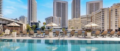 Un espace aménagé au bord de la piscine, avec des chaises longues et des parasols, entouré de grands immeubles.