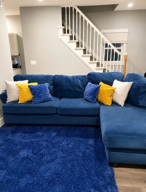 A blue sectional sofa with yellow and white cushions, a blue shag rug, and a staircase with white railings.