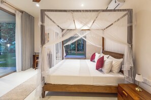 A four-poster canopy bed with white bedding and red pillows, a wooden nightstand, and a view of a garden through a large window. - ONA Garden Villa: Deluxe Double Room (Galle)