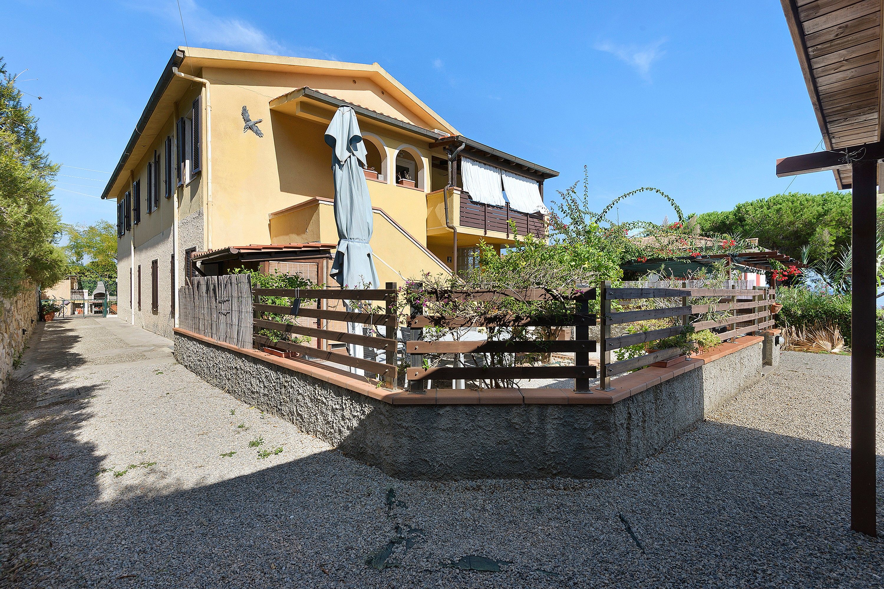 A yellow building with a balcony, a wooden fence, and a gravel path.