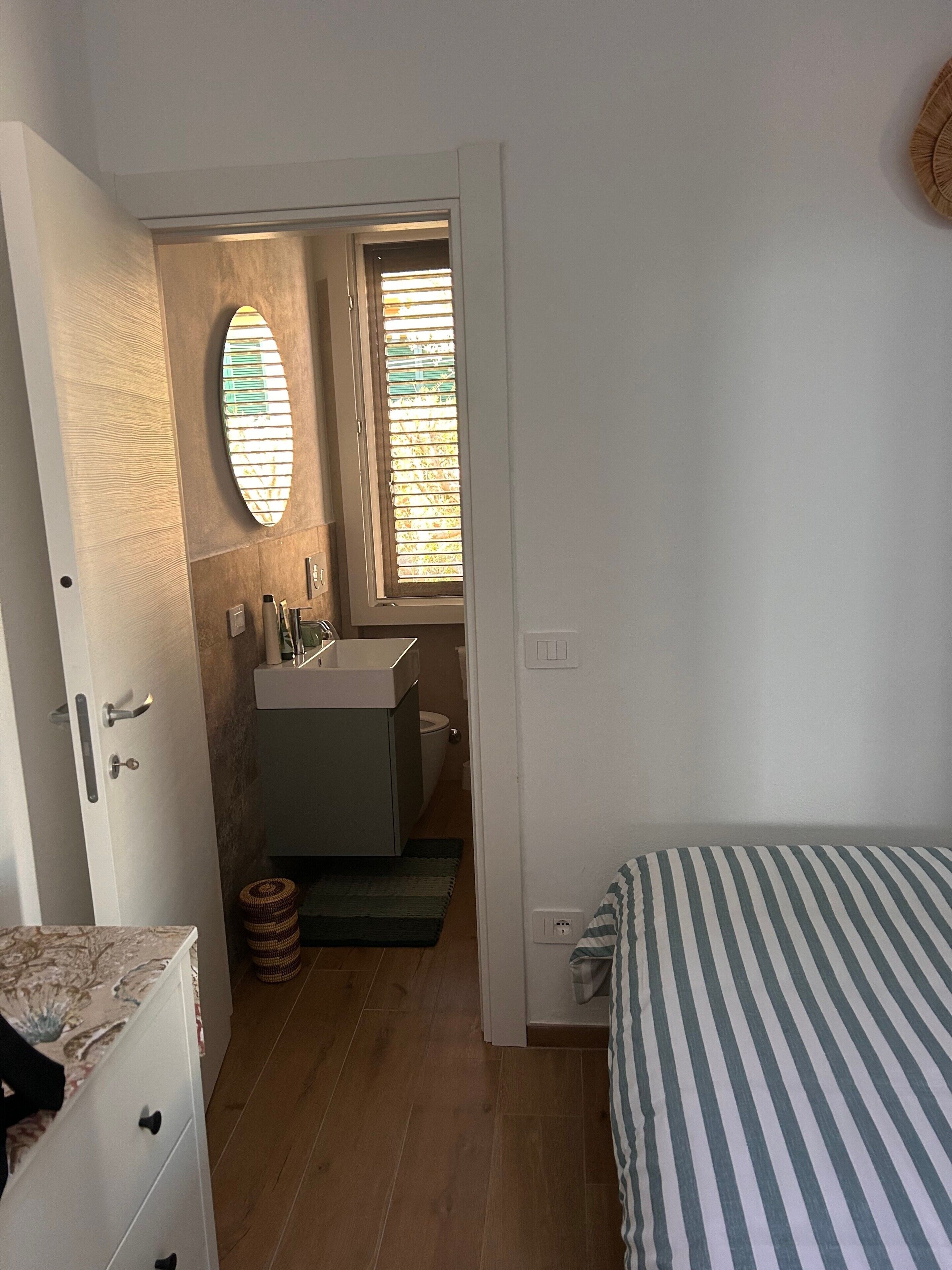 Un dormitorio con cama, cómoda, tocador con espejo, ventana con persianas y puerta que da a otra habitación.
