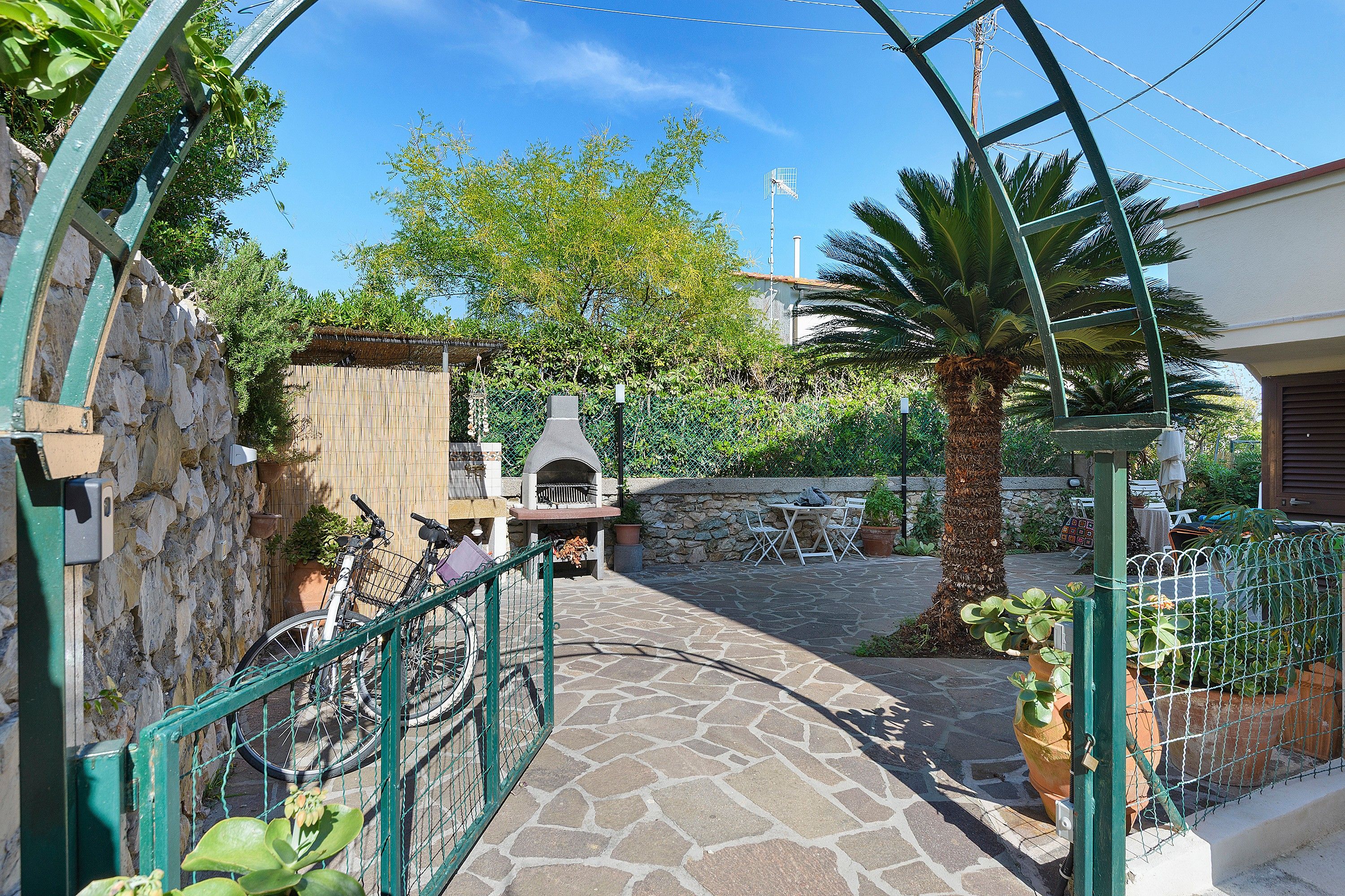A stone pathway leading to a courtyard with a barbecue area, a bicycle, and a palm tree.