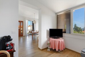 A room with a television on a small table, a dining area with chairs and a table, and a window with blinds.