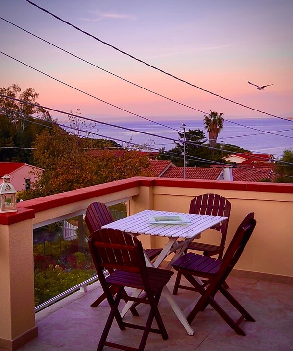 A balcony with a table and chairs, overlooking a residential area and the sea during sunset.