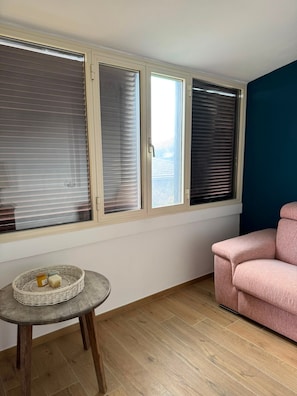 A room with a wooden floor, a round table with a basket, a pink sofa, and three windows with blinds.