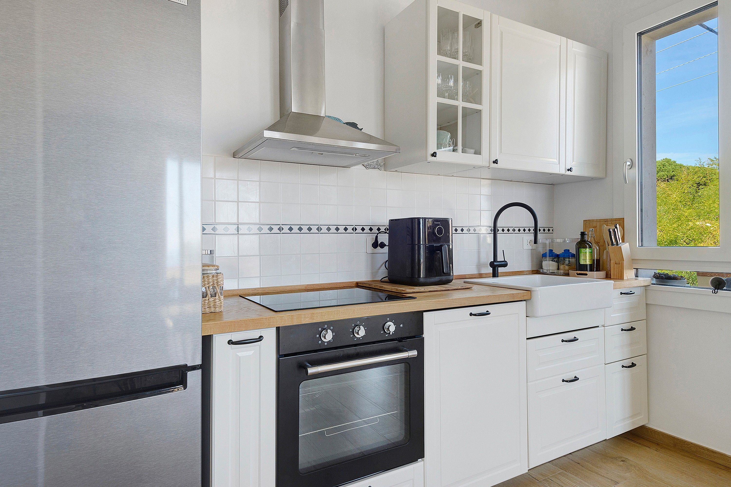 Eine moderne Küche mit weißen Schränken, einem Edelstahlkühlschrank, einem eingebauten Backofen und einem Fenster mit Blick auf Grünflächen.