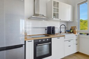 A modern kitchen with white cabinetry, a stainless steel refrigerator, a built-in oven, and a window providing a view of greenery outside.