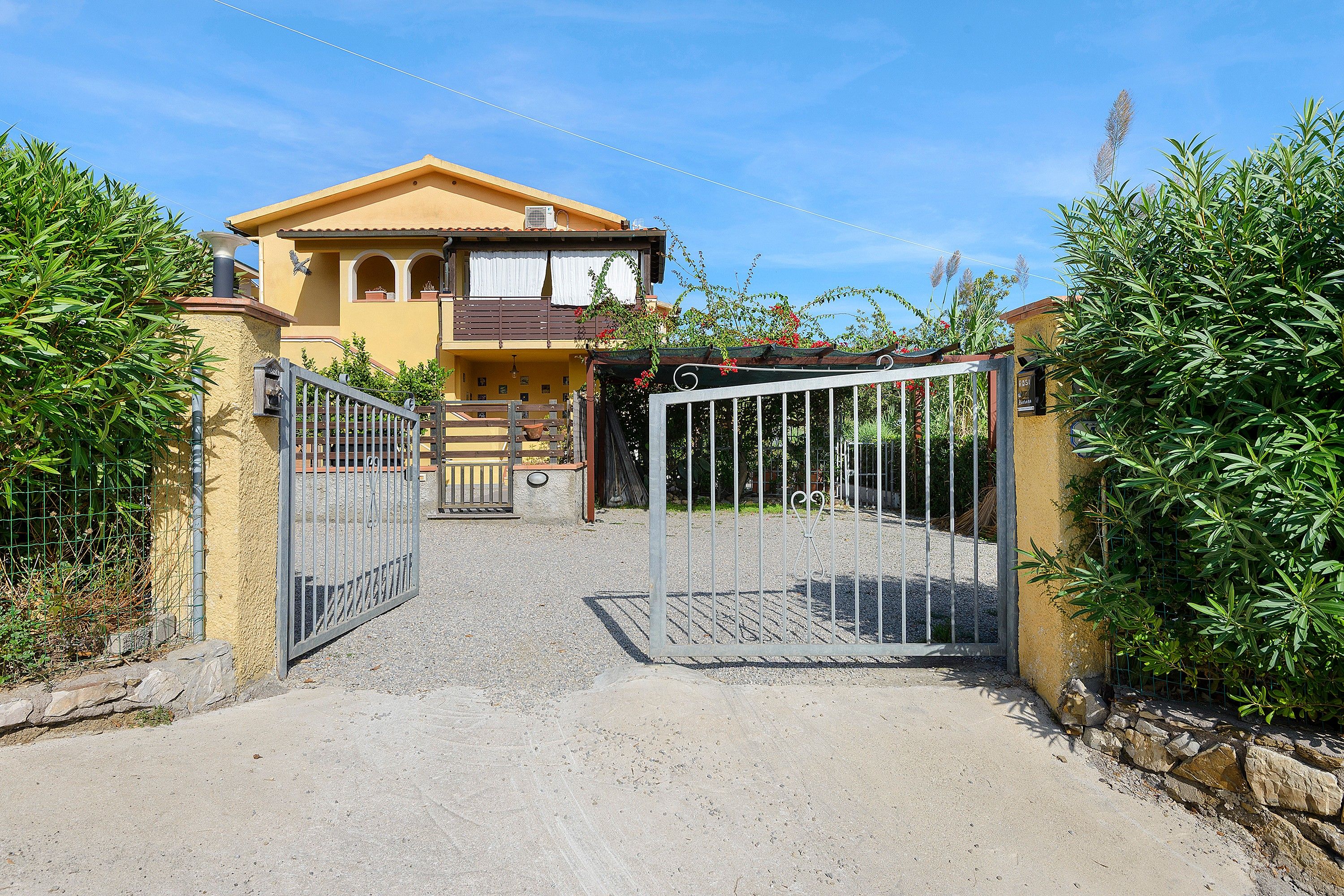 A gated entrance to a property with a yellow facade, a balcony with a railing, and a gravel driveway.