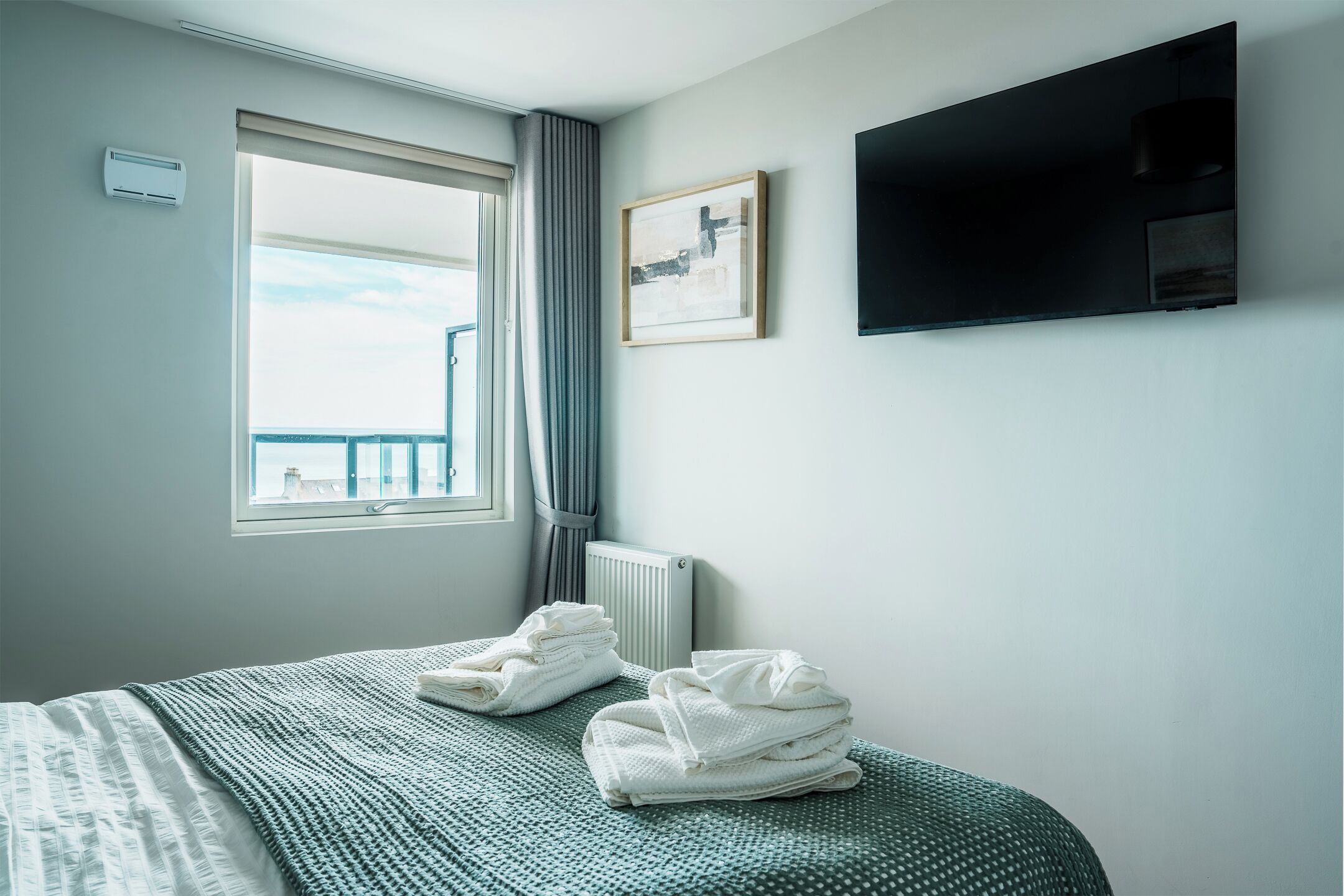 Quarto de hotel com cama, radiador, janela com cortinas, quadro na parede e televisão de tela plana.