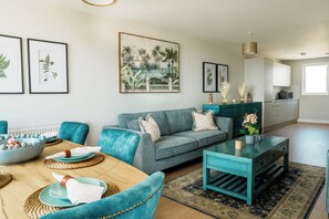 A modern living room with a teal sofa, a dining table with plates and a bowl, and a kitchen area with cabinets and a window.