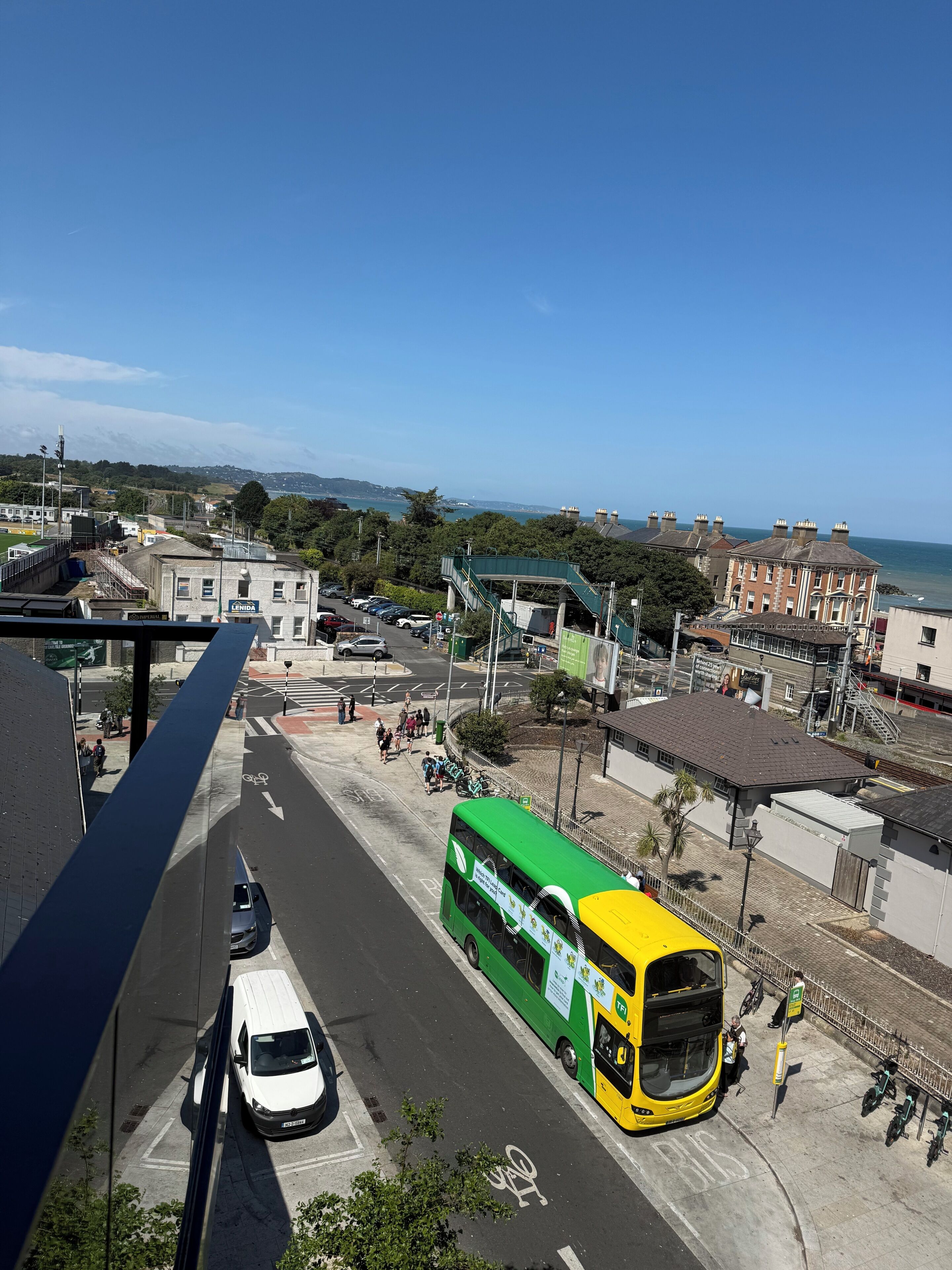 A green and yellow bus at a bus stop with a clear view of the sea and buildings in the background.