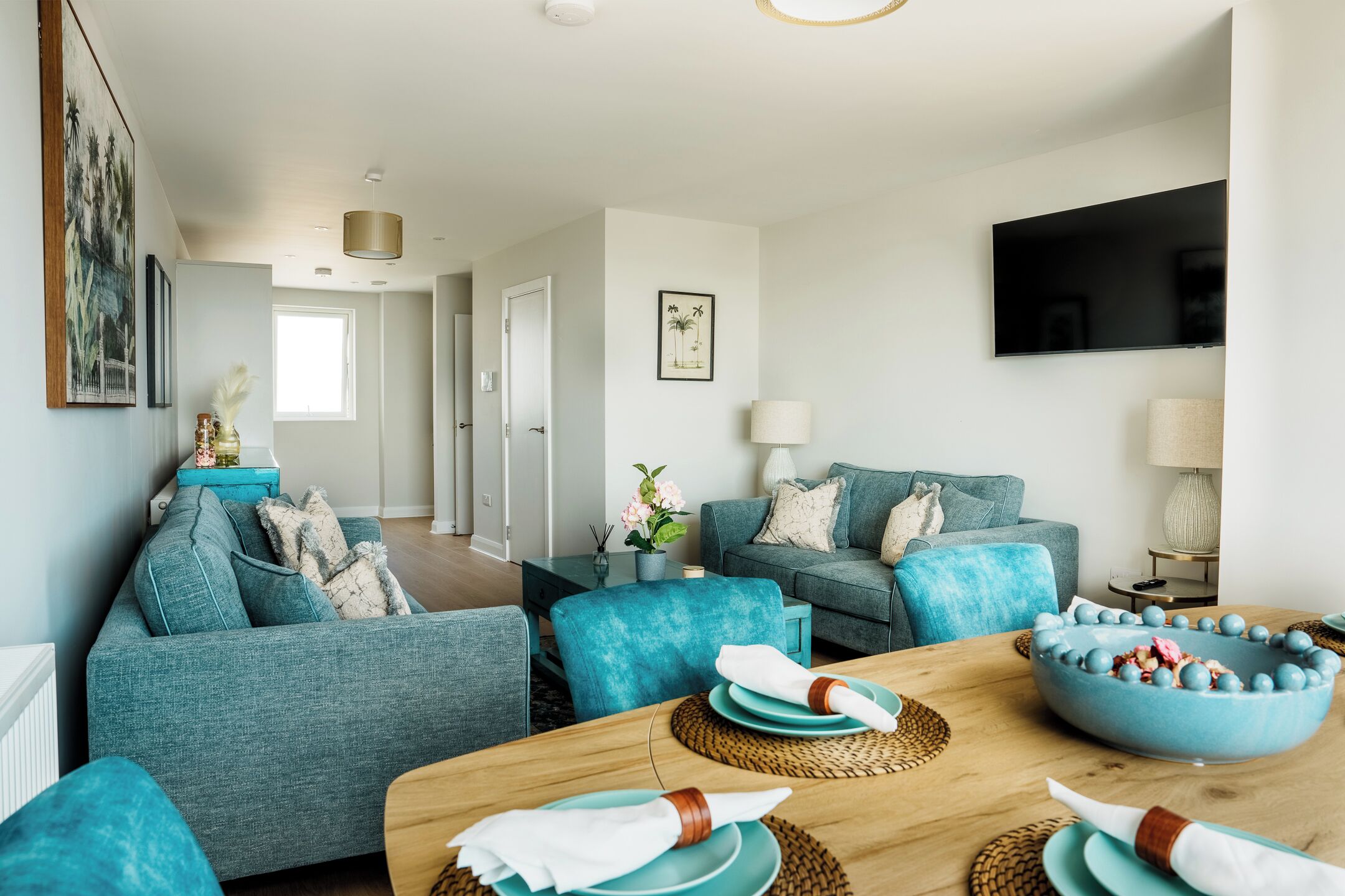A modern living room with teal sofas, a wooden dining table, and a flat-screen TV mounted on the wall.