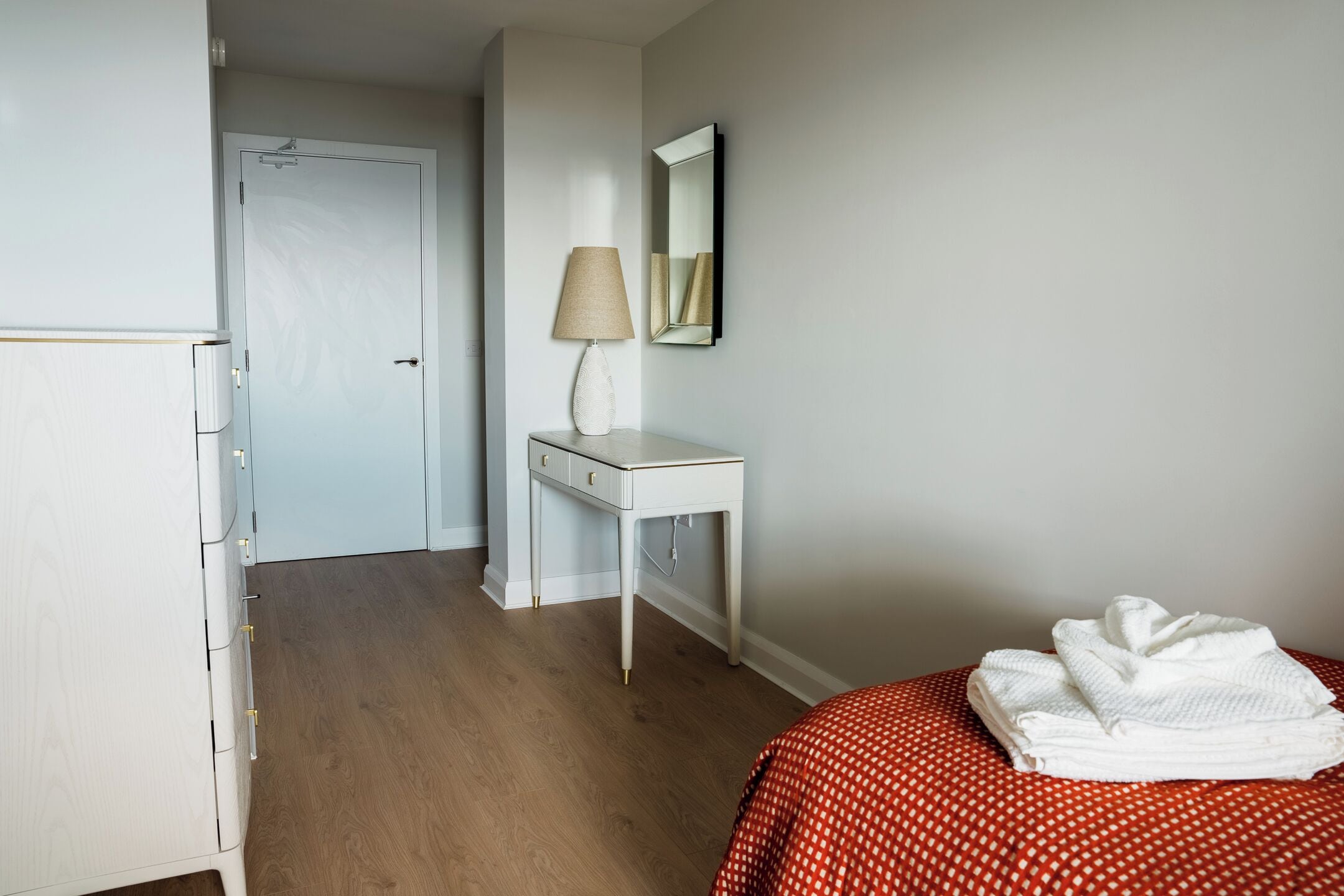 Ein Hotelzimmer mit Holzboden, einem weißen Kleiderschrank, einem Nachttisch mit Lampe, einem Spiegel und einem Bett mit rot-weißer Bettdecke.