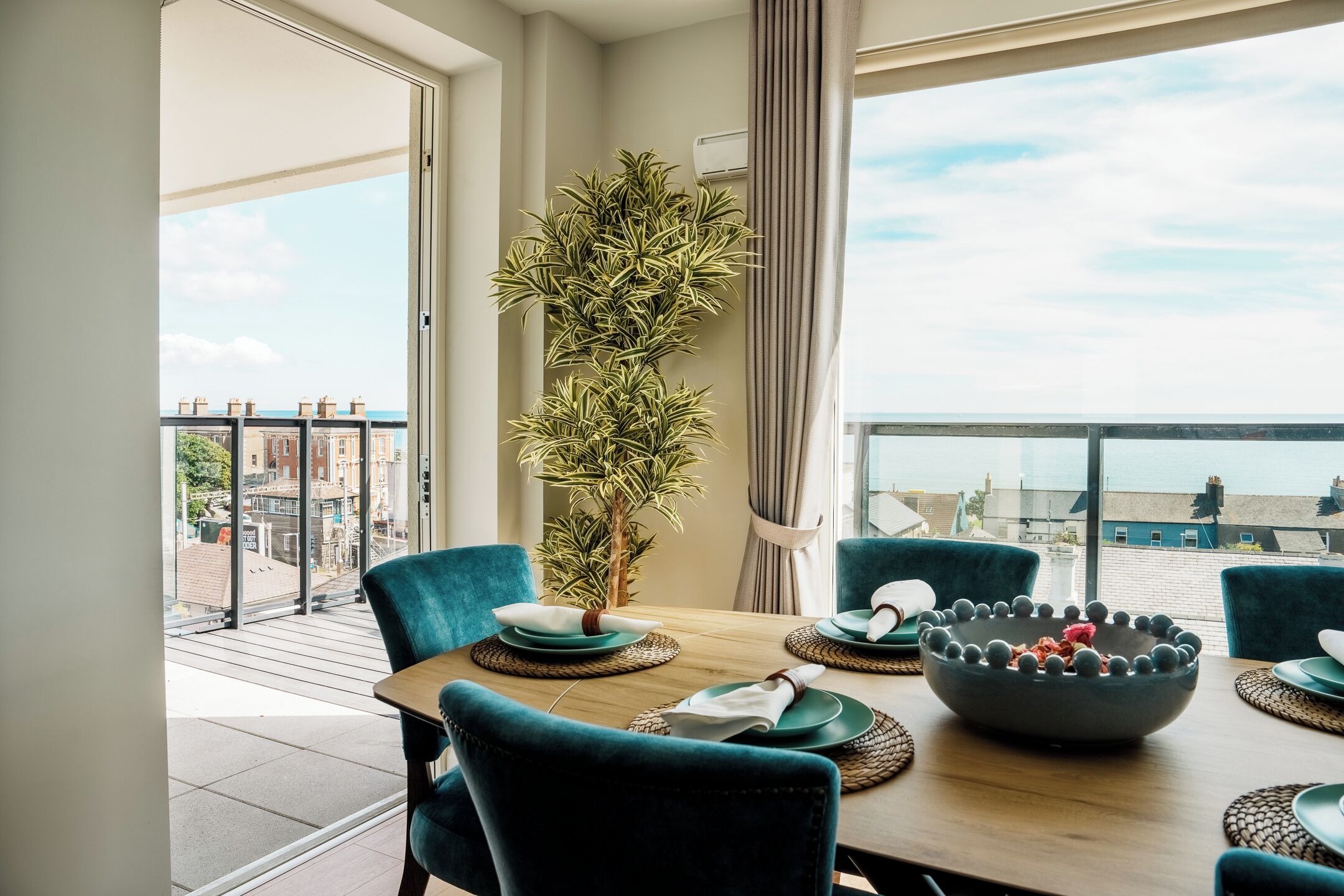 Un comedor con una mesa dispuesta para cuatro, una planta en maceta y vistas de la ciudad a través de grandes ventanales.