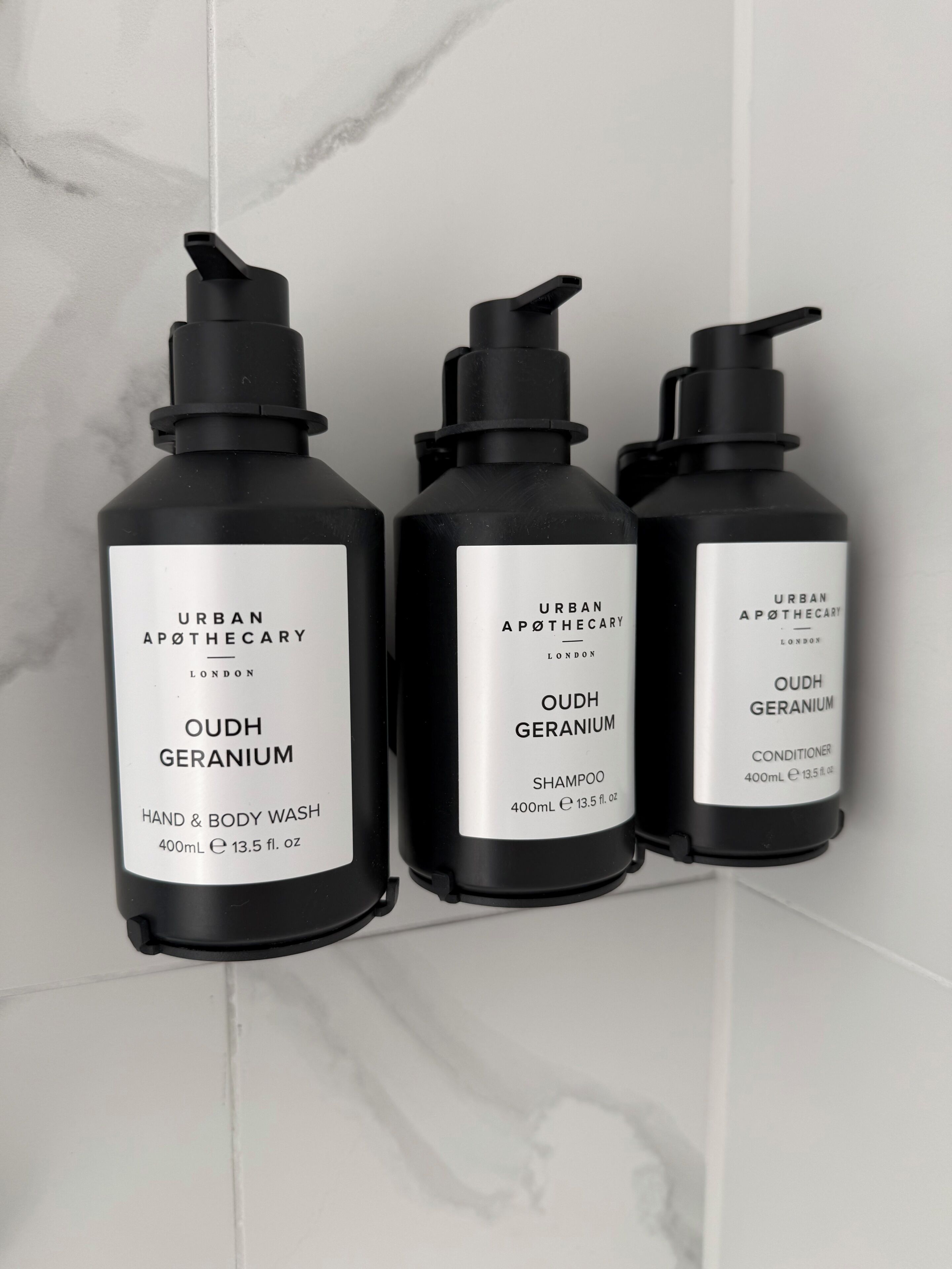 Three black bottles of Oudh Geranium products on a white tiled wall.