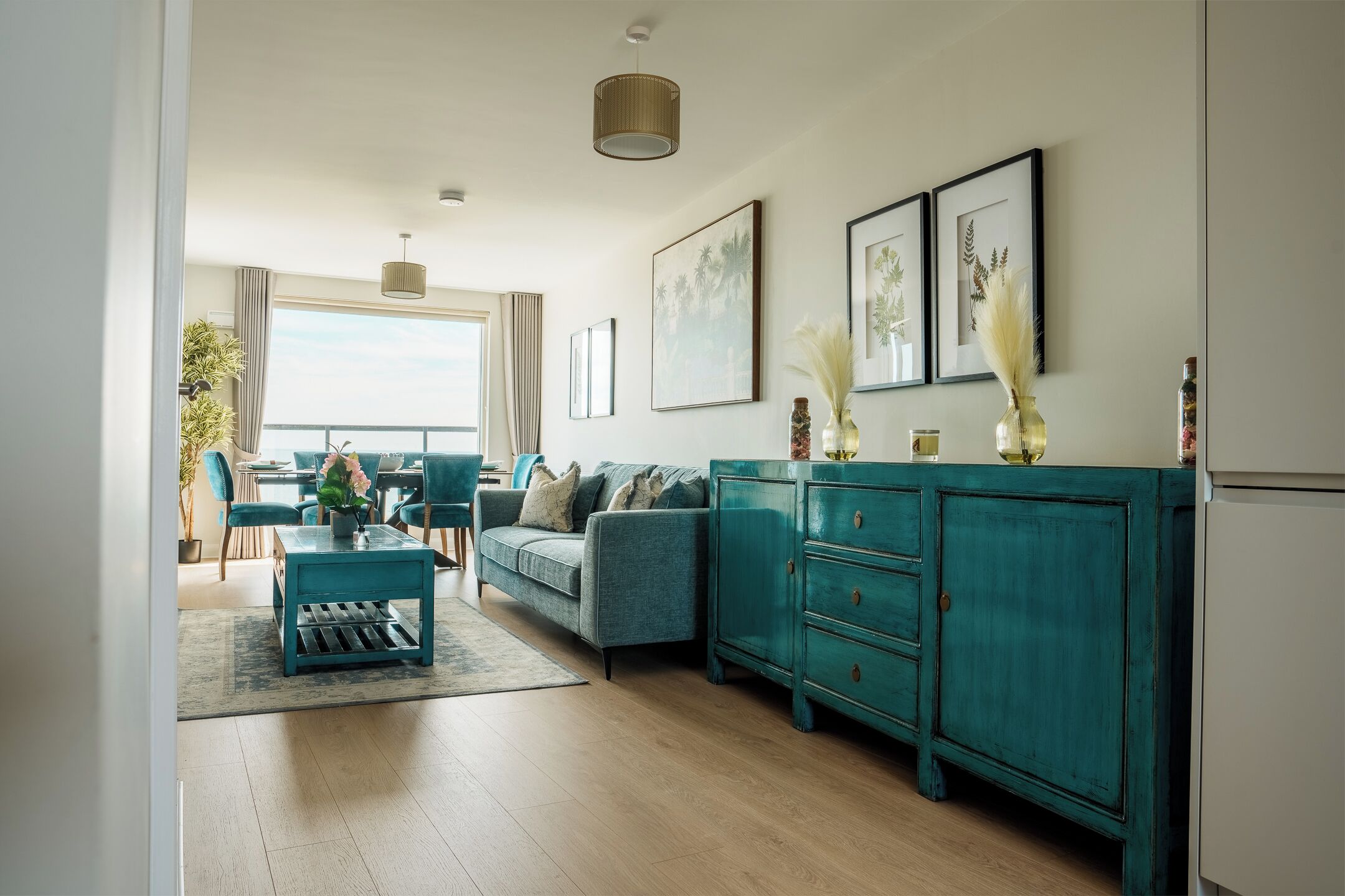 Een moderne woonkamer met een blauwgroene bank, een salontafel en een kast. Er hangen ingelijste foto's aan de muur en staat een plant op het raamkozijn.
