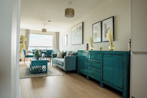 A modern living room with a teal sofa, a coffee table, and a cabinet. There are framed pictures on the wall and a plant on the windowsill.