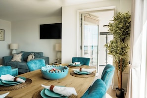 A modern living room with a teal sofa, a dining table with blue plates, and a large plant.
