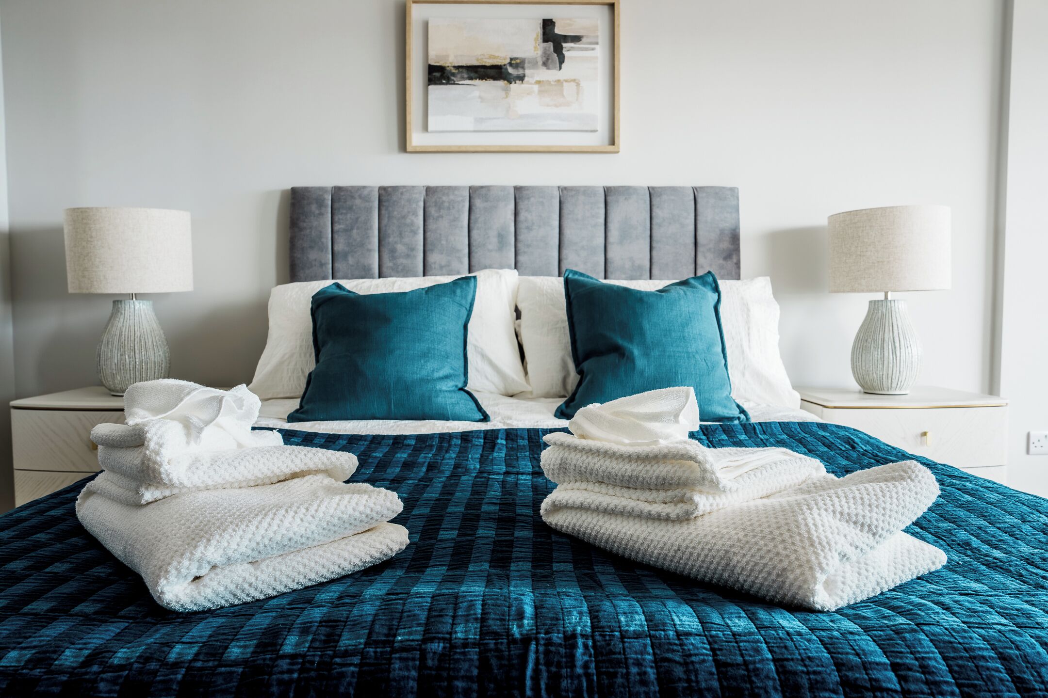 Cama bem arrumada com colcha azul, duas toalhas brancas e almofadas verde-água.