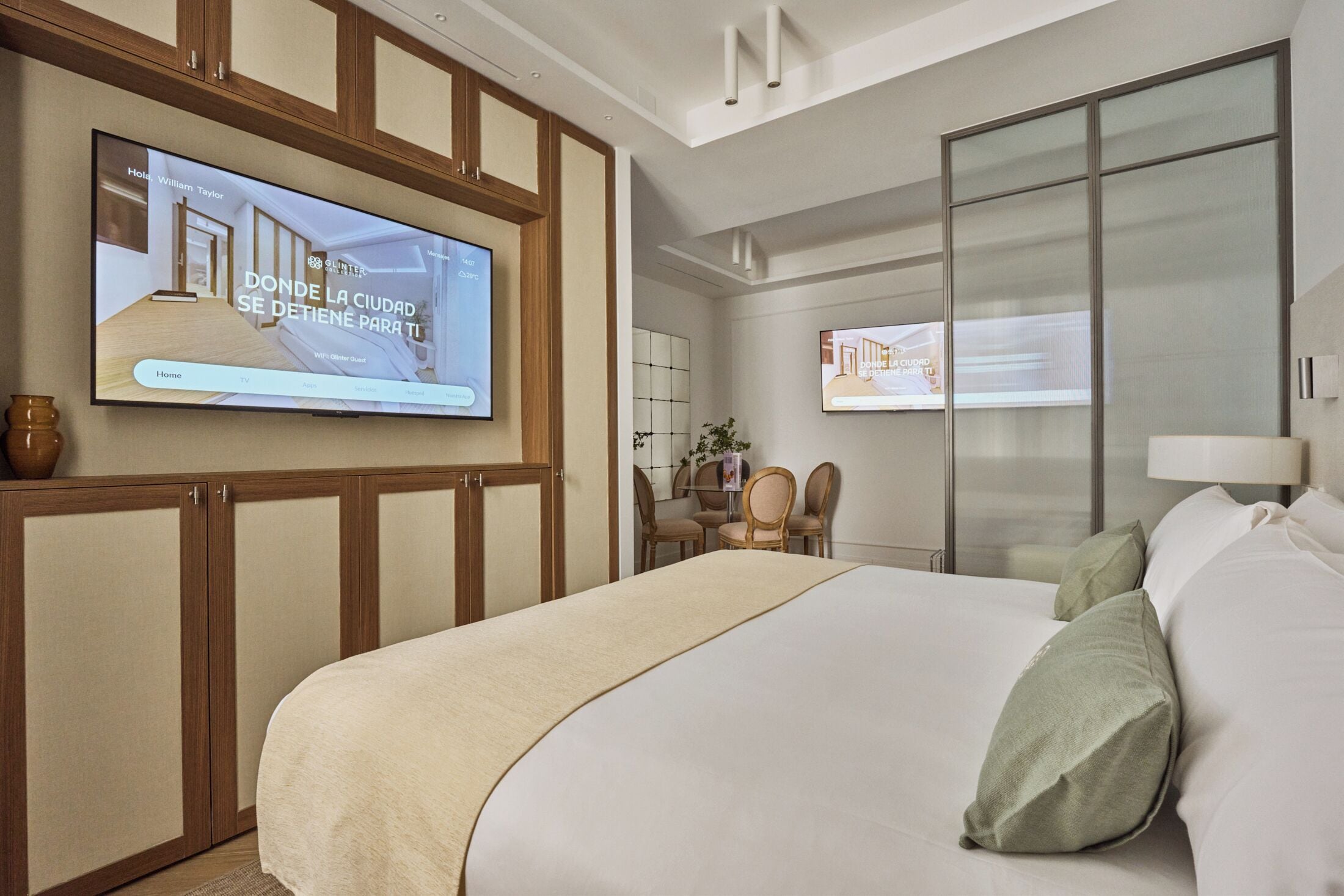 Ein Hotelzimmer mit einem großen Bett, einem an der Wand befestigten Fernseher, einem Essbereich mit Stühlen und einer Schiebetür aus Glas.