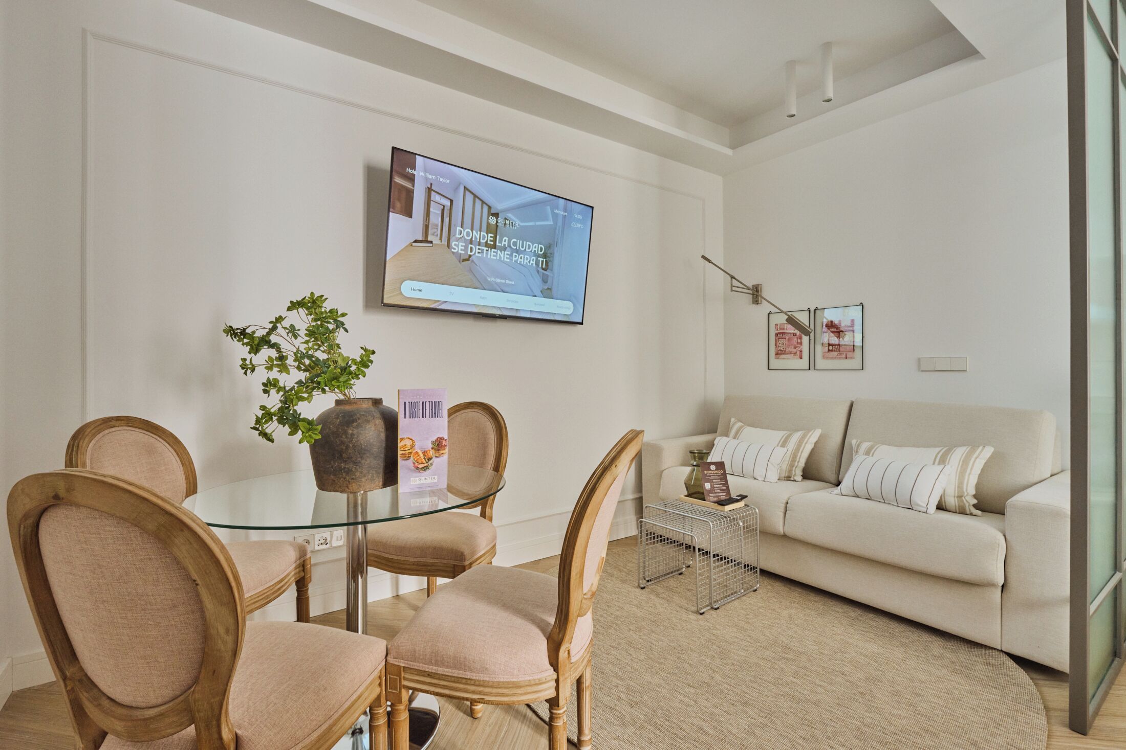 Un salón moderno con una mesa de comedor de vidrio, dos sillas de madera, un sofá blanco y un televisor montado en la pared.
