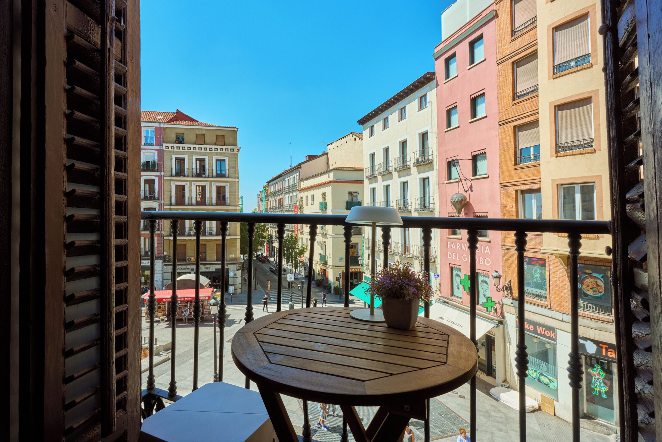 Un balcón con mesa de madera y una planta en maceta, con vista a una calle con edificios y comercios.