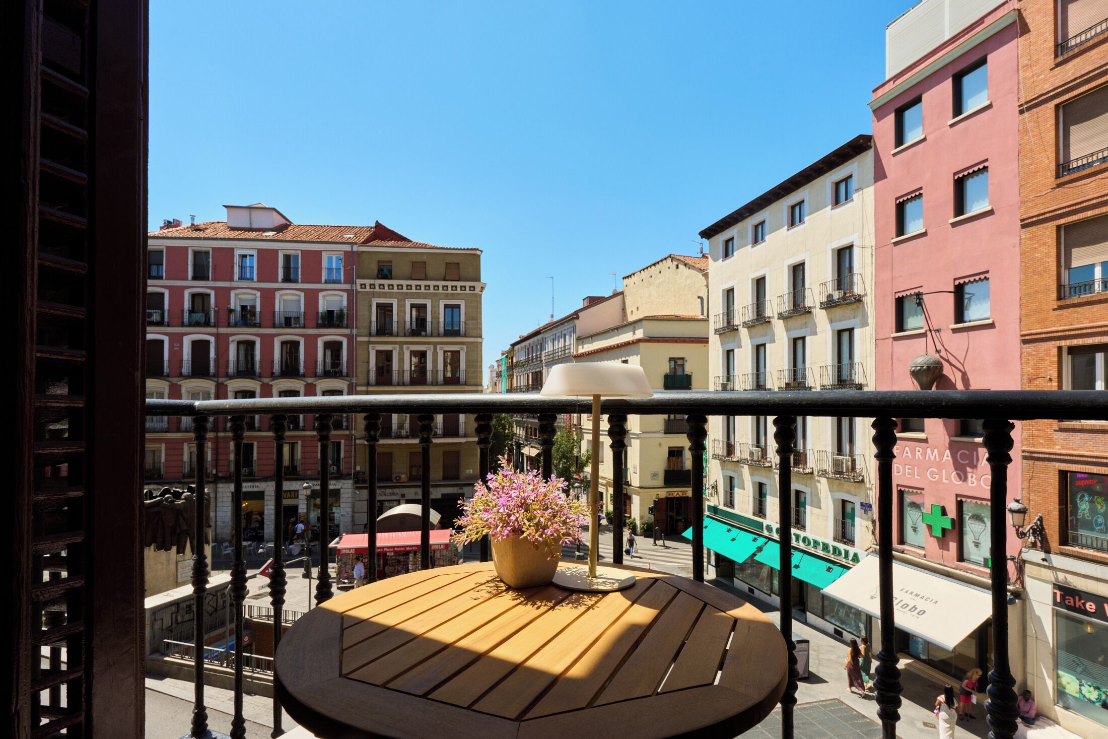 A balcony with a wooden table and a potted plant, overlooking a street with buildings and shops.