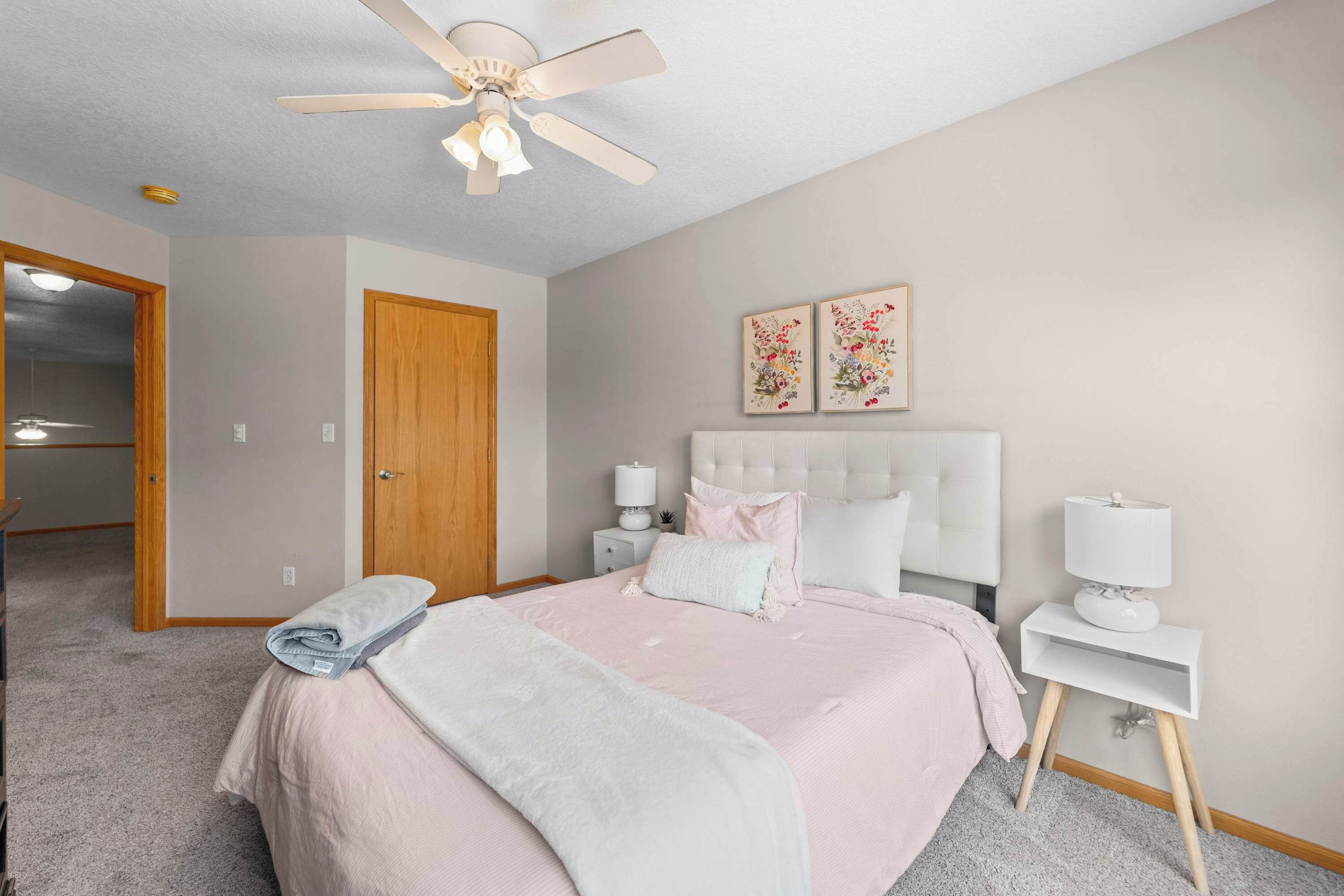 Une chambre à coucher comprenant un lit, une table de chevet, un ventilateur de plafond, une porte et deux œuvres d’art mural.