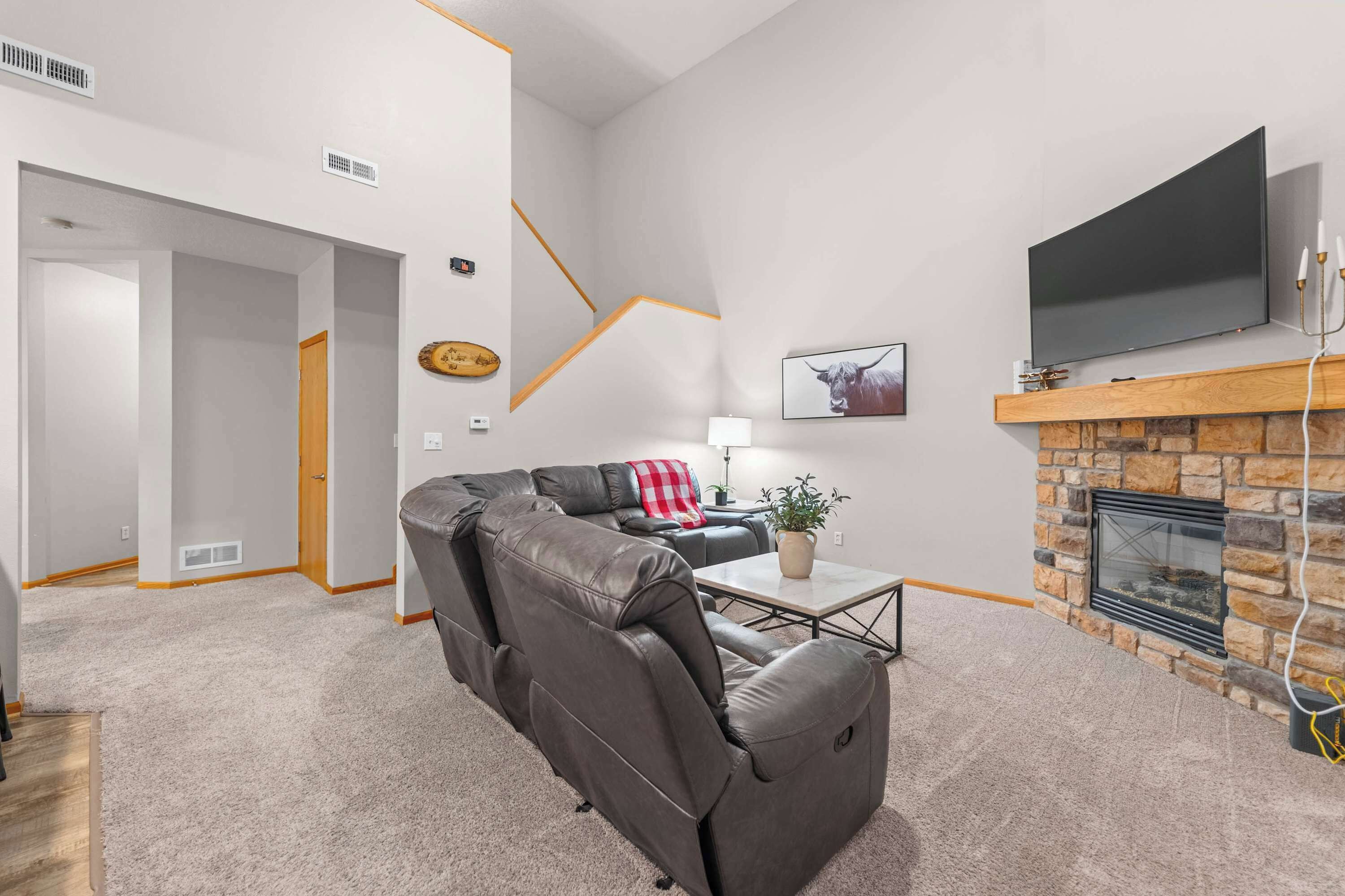 A modern living room with a leather sofa, a flat-screen TV mounted above a stone fireplace, and a hallway leading to another room.