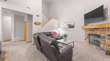 A modern living room with a leather sofa, a flat-screen TV mounted above a stone fireplace, and a hallway leading to another room.